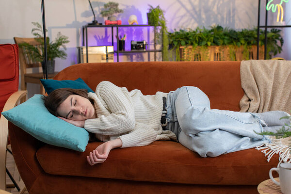 Tired young woman lying down in bed taking a rest at home apartment room. Carefree pretty girl napping, falling asleep on comfortable orange sofa with pillows. Closed her eyes enjoy daytime nap alone