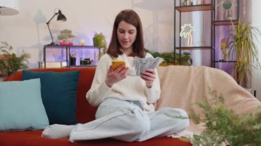 Aile bütçesi planlamak. Gülümseyen genç bir kadın nakit para sayıyor, akıllı telefon kullanıyor ev faturalarını hesaplıyor. Neşeli kız gelirinden memnun ve planlı tatil ve hediyeler için para biriktiriyor.