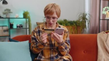 Woman using credit bank card and smartphone while transferring money, purchases online shopping cashless, order food delivery at home apartment indoors. Happy young girl sitting in room on couch