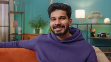 Portrait of happy calm indian man at home couch smiling friendly, glad expression looking away dreaming resting, relaxation feel satisfied concept good news, celebrate win. Hindu guy in living room