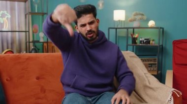 Dislike. Upset indian man showing thumbs down sign gesture, expressing discontent, disapproval dissatisfied bad work at modern home apartment indoors. Displeased hindu guy in living room on sofa