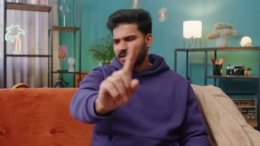 Memnun olmayan Hintli adam nahoş berbat bir fikre tepki veriyor, kötü kaliteden memnun değil, el sallıyor, başını sallıyor hayır, reddet fikri, evlenme teklifini sevme. Evdeki kanepede oturan Hintli genç adam.