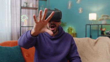 Indian man using virtual reality futuristic technology VR app headset helmet to play simulation 3D 360 video game, watching film movie at modern home apartment. Hindu guy in goggles sitting on couch