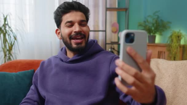 幸せなインドの男性ブロガーは スマートフォンで自撮りを取り ソーシャルメディアの購読者とオンラインでビデオ通話を通信し ブログ記事を記録します 若いですHindu男で家のアパートの部屋オンソファ — ストック動画