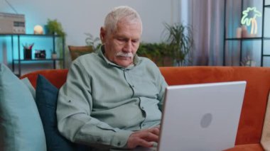 Büyükbabam evdeki kanepede oturduktan sonra dizüstü bilgisayarını kapatıyor. Çevrimiçi çalışan yaşlı bir adam, uzaktan kumanda. Defter bilgisayarında e-öğrenme, internette gezinme