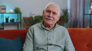 Mutlu, sakin, yaşlı büyükbaba kanepede gülümseyen dostça gülümseyen, mutlu bir yüz ifadesi olan dinlenme, rahatlama konsepti iyi haber, zaferi kutlayın. Oturma odasındaki yaşlı adam.