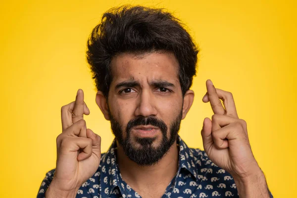 Indian young man crossed fingers looking at camera asking for good lucky news, wishing good exam results, dreaming about win victory in lottery jackpot. Hindu guy isolated on yellow studio background