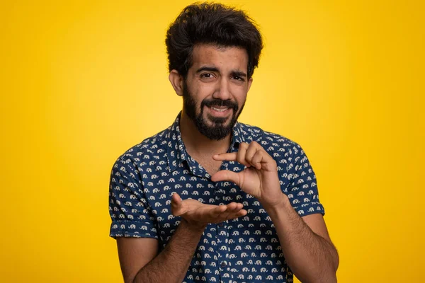 Need some more, please give me. Indian man showing a little bit gesture with sceptic smile, showing minimum sign, measuring small size. Handsome hindu guy isolated alone on yellow studio background