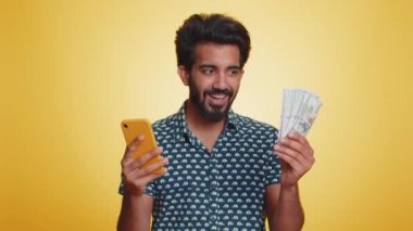 Akıllı telefona bakan Hintli bir adam içten bir sevinç gösterisi kazanır, para dolar banknotları, büyük ikramiye başarısı, gelir maaşı alır. Hintli adam sarı arka planda izole edilmiş.