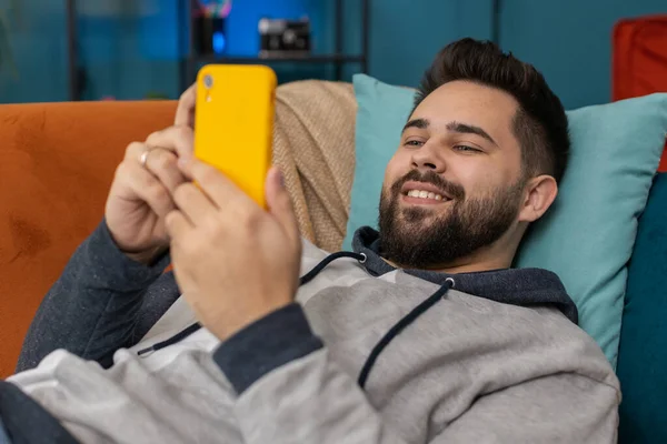 Happy man lying on couch uses mobile phone smiles at home living room apartment. Caucasian guy texting share messages content on smartphone social media applications online, watching relax movie