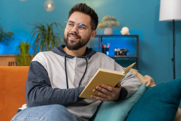 Young man relaxing interesting book, turning pages smiling enjoying literature, taking a rest on comfortable couch. Portrait of peaceful cheerful Caucasian guy at home apartment living room on sofa
