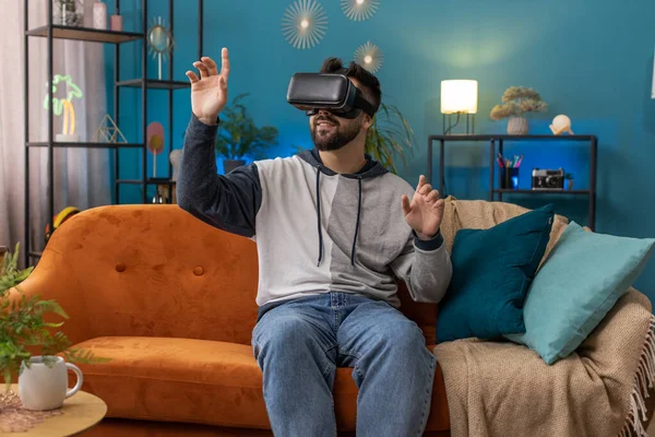 Excited man using virtual reality futuristic technology VR app headset helmet to play simulation 3D 360 video game watching film movie. Portrait of guy in goggles at modern home apartment room on sofa