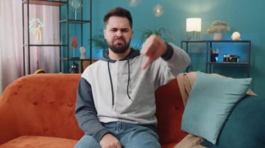 Hoşlanmamak. Sinirli genç adam işaret dilini gösteriyor, hoşnutsuzluğunu dile getiriyor, hoşnutsuzluğunu dile getiriyor, modern apartman dairesinde kötü işler yapıyor. Oturma odasındaki kanepede oturan beyaz adam.