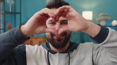 Ben de seni seviyorum. Oturma odasındaki kanepede oturan mutlu beyaz adam sevgiyi simgeliyor. Kameraya kalp işaretini gösteriyor. Romantik duygular samimi olumlu duygular ifade ediyor. Sadaka, minnettarlık, bağış.