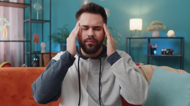 不満を持った男が寺院をこすり 頭痛の種を治すために 緊張と片頭痛に苦しみ 自宅の部屋のアパートで痛みの高血圧でストレスの痛みをつかむ ソファの上の白人男性 ライフスタイル — ストック動画