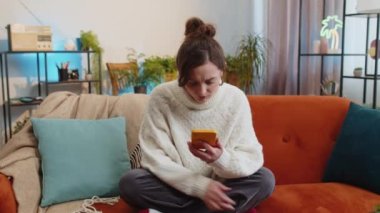 Hüzünlü kadın akıllı telefon daktilo kullanıyor, beklenmedik piyango sonuçlarını kaybediyor, kötü haber, servet kaybı, başarısızlık, teslim tarihi, virüs. Evdeki kanepede oturan genç kızın portresi.