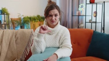 Dislike. Portrait of upset woman showing thumbs down sign gesture, expressing discontent, disapproval dissatisfied bad work at modern home apartment indoors. Displeased girl in living room on sofa