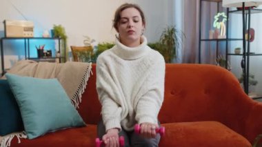 Kadın halterde ağırlık kaldırıyor, evde pazı kaldırıyor. Egzersiz, yağlı kalori yakma, antrenman gücü, ev içi spor aerobiği. Spor motivasyonu. Kanepede oturan esmer kız.