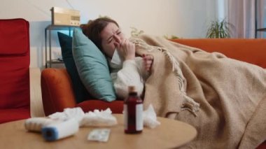 Evdeki oturma odasında kanepede oturan soğuk algınlığı ya da alerjiden muzdarip genç hasta kadın. Hasta kız ekose darbelere sarılı şapka giyiyor hapşırıyor sümük peçeteye yapışıyor. Coronavirus karantinası, salgın