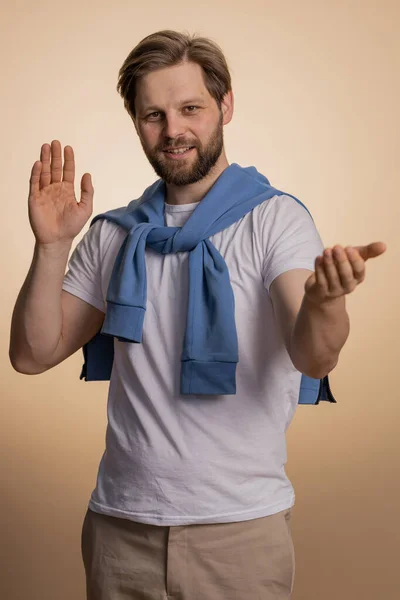Come here, join us. Welcome. Young bearded man showing inviting gesture with hands, ask to join, beckoning to coming, gesturing hello, goodbye. Caucasian guy isolated on beige studio background