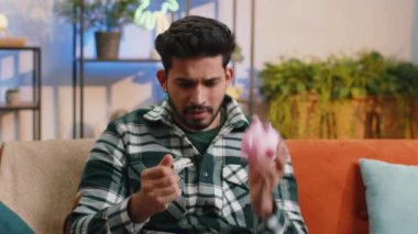 Zavallı Hintli genç adam yetersiz miktarda para elinde kumbara ve bir dolarlık banknot tutuyor. Finansal kriz. İflas. Yoksulluk ve yoksulluk. Hintli adam kanepede oturuyor.