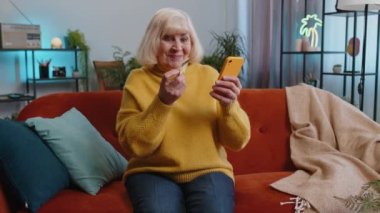 Büyük anne kredi kartı ve akıllı telefon kullanarak para transfer ediyor, çevrimiçi alışveriş nakitsiz, yemek siparişi veriyor, ev içinde faturaları ödüyor. Kanepedeki yaşlı kadın.