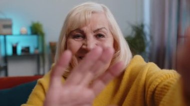 Üst düzey büyükanne kadının internetten selfie videosu çekerken el sallayarak ev içinde kameraya baktığı görüntüler. İletişim, toplantı. Yaşlı blogcu akıllı telefon konuşması yapıyor