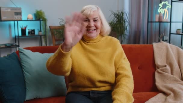 こんにちは シニア女性のカメラでフレンドリーな笑みを浮かべて 手こんにちは 挨拶や別れをジェスチャー手を振って 自宅でもてなしの表現を歓迎する おばあちゃんリビングでソファに座って — ストック動画