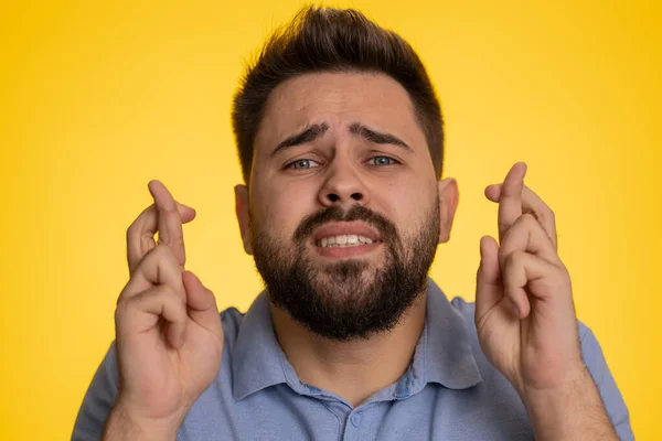 Young man crossed fingers looking at camera asking for good luck news, wishing good education exam results, dreaming about win victory in lottery jackpot. Guy isolated on yellow studio background