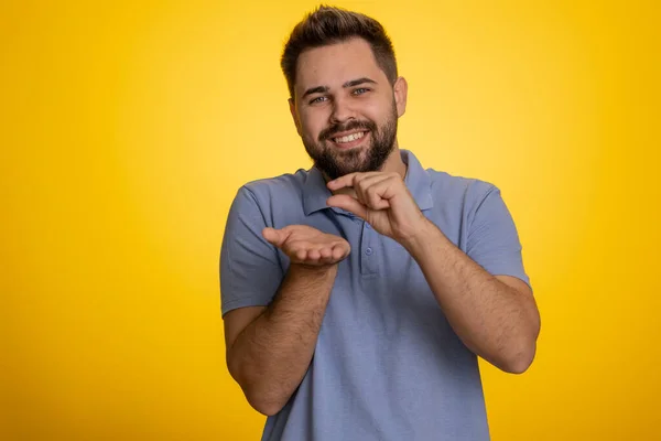 Need some more, please give me. Caucasian man showing a little bit gesture with sceptic smile, showing minimum sign, measuring small size. Handsome guy isolated alone on yellow studio background