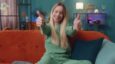 Gibi. Mutlu, heyecanlı, beyaz bir kadın kameraya bakıp olumlu bir şey işaret ediyor. İyi haber, olumlu geri bildirim gibi. Oturma odasındaki kanepede oturan genç kız.