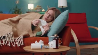 Evdeki oturma odasındaki kanepede yatan soğuk algınlığı ya da alerjiden muzdarip genç hasta kadın. Kafasında havlu olan hasta çocuk hapşırır, sümüğü peçeteye sürer. Coronavirus karantina salgını
