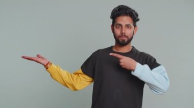 Mutlu Hintli genç adam başparmaklarını gösterip boş yeri işaret ediyor. Ticari metin yazıları için reklam alanı, ürün tanıtım afişi için fotokopi alanı. Hintli adam gri stüdyo arka planında izole edilmiş.