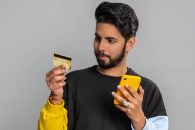 Para transferi, online alışveriş, ödeme yaparken kredi kartı ve akıllı telefon kullanan samimi Hintli bir müşteri. Finans ve internet. Yakışıklı adam gri arka planda tek başına.