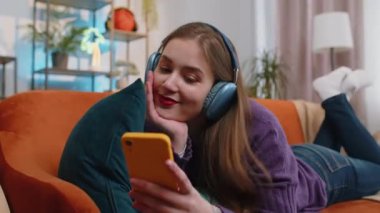 Kablosuz kulaklıklı mutlu genç bir kadın evinde koltukta uzanmış en sevilen enerjik disko dans müziğini dinliyor. İnsanlar hafta sonu boş zaman aktiviteleri