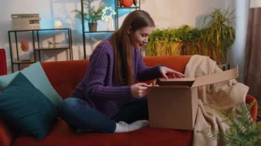 Evdeki kanepede oturan mutlu, genç bir kadın teslimat paketini açıyor. Gülümseyen tatmin olmuş kız alışverişci internet müşterisi karton kutu açıyor hızlı posta yoluyla satın alma hediyesi alıyor