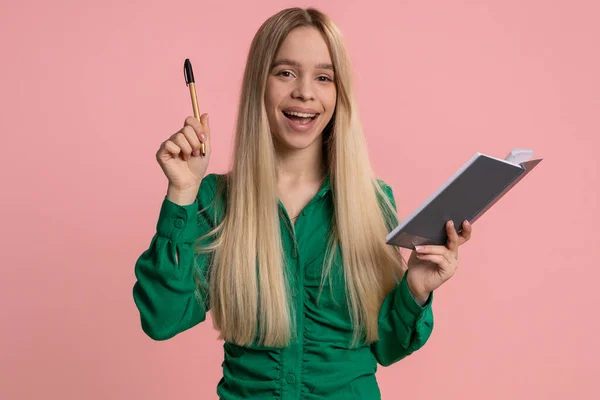 Thoughtful journalist caucasian woman making notes, writing down thoughts with pen into notepad notebook diary, to do list, good idea. Pretty teenager girl isolated alone on pink studio background