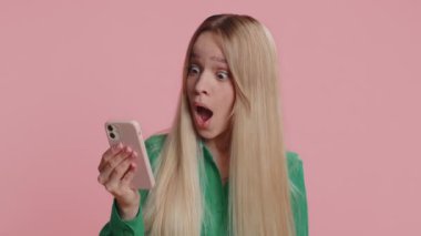Akıllı telefonlara bakan mutlu bir kadın samimi bir şekilde sevinçle zaferi kutluyor, para dolu banknotlar alıyor, büyük ikramiye başarısı elde ediyor. Genç kız pembe stüdyo geçmişinde izole edilmiş.
