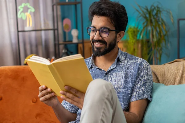 若いインド人の男は面白い本をリラックスし ページをめくる文学を楽しんで笑顔 快適なソファの上で休息を取る ポートレートの平和な陽気Hindu男にホームアパートリビングルームでソファ — ストック写真