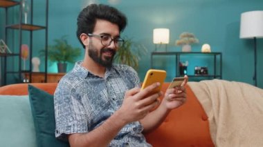 Hintli bir adam parayı transfer ederken kredi kartı ve akıllı telefon kullanıyor. İnternetten nakitsiz alışveriş yapıyor. Ev içinde yemek siparişi veriyor. Koltukta oturan mutlu Hintli adam.