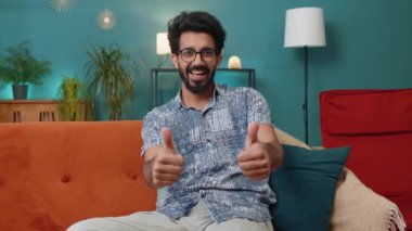 Gibi. Mutlu heyecanlı Hintli adam onaylayıcı bir şekilde kameraya bakıyor olumlu bir şey imzalıyor, iyi haber, olumlu geri bildirim gibi. Oturma odasındaki kanepede oturan genç Hintli.