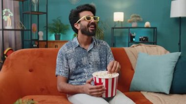 Excited indian man in 3D glasses sits on couch eating popcorn snacks and watching interesting TV serial sport game, film online social media movie content at home apartment. Guy enjoying entertainment