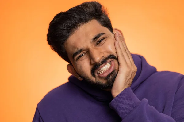Indian bearded man touching sore cheek suffering from toothache cavities, gingivitis waiting for dentist appointment gums disease. Handsome hindu guy indoors studio shot isolated on orange background