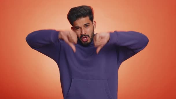 嫌いだ 嫌いを表現し 親指を看板ジェスチャーを示すパーカーで不幸なインド人の男を動揺 ハンサムな若い男 オレンジを背景に撮影室内スタジオ — ストック動画