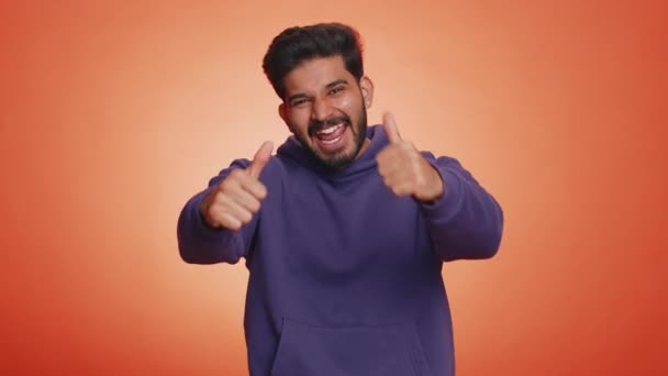 まるで パーカーのインドの若い男は親指を上げる何かに同意するか 肯定的な応答を与える良いような広告をお勧めします ハンサムな髭の後ろ男隔離上のオレンジ色のスタジオの背景 — ストック動画