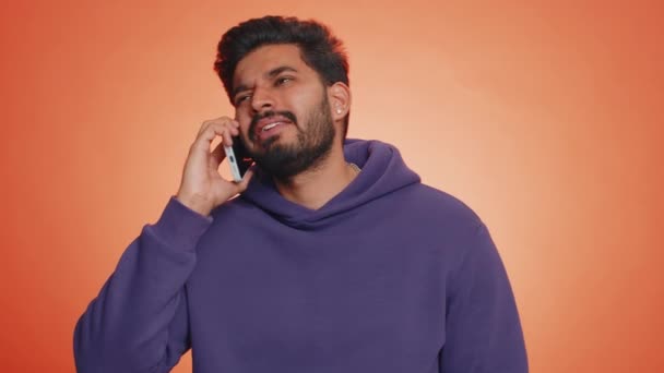 若い疲れ眠いインドのフリーランスの男は友人と携帯電話で話し オンライン会話をしています 興味のないHindu男を悩ませています退屈な話上のスマートフォン隔離されたオンオレンジ背景 — ストック動画