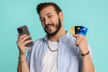 Para transferi, online alışveriş, ödeme yaparken kredi kartı ve akıllı telefon kullanan Lübnanlı samimi müşteri. Finans ve internet. Yakışıklı adam mavi arka planda tek başına.