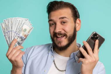 Lübnanlı adam, akıllı telefon görünümlü, neşeli bir galibiyet sergiliyor. Nakit dolar banknotları alıyor. Piyango talihi, gelir maaşı. Stüdyo mavi arka planda izole edilmiş Arap bir adam.