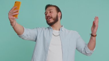 Lübnanlı gezgin blogcu, akıllı telefondan selfie çekiyor, aile veya arkadaşlarla çevrimiçi video görüşmesi yapıyor, canlı çeviri akışı yapıyor. Yakışıklı Arap adam mavi arka planda, kapalı alanda izole edilmiş.
