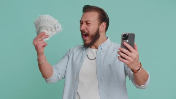 レバノン人の男性はスマートフォンを探して誠実に勝利を喜び お金を受け取るドルの現金紙幣 成功宝くじのジャックポット運 所得給与の賃金を表示します スタジオブルーの背景に隔離されたアラビア人 — ストック動画